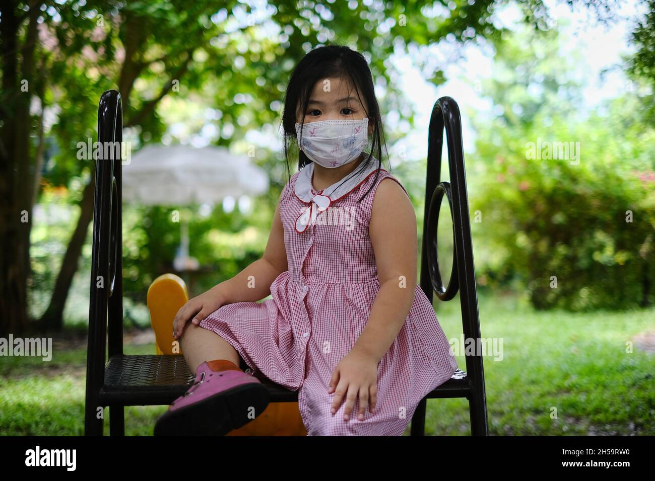 A cute young chubby Asian girl in red and white dress is sitting at the top of a slide in a park, resting. Kid with face mask playing outdoor alone du Stock Photo