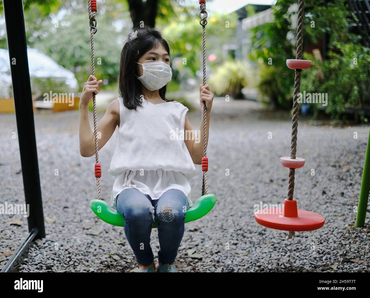 A cute young Asian girl with a white face mask is playing alone on a swing in a playground during the Covid-19 pandemic. Stock Photo