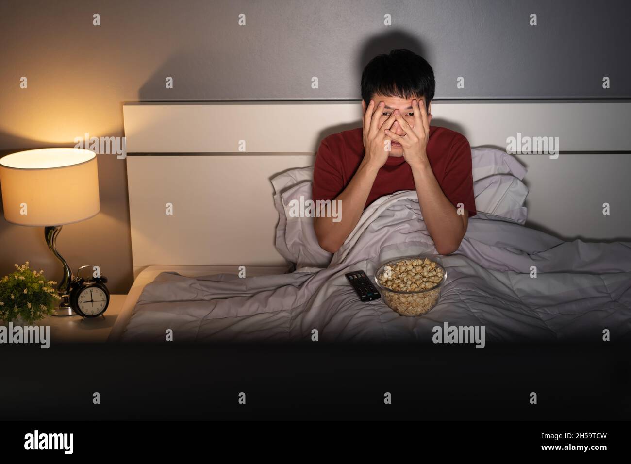 scared young man is watching horror movie TV on a bed at night Stock Photo