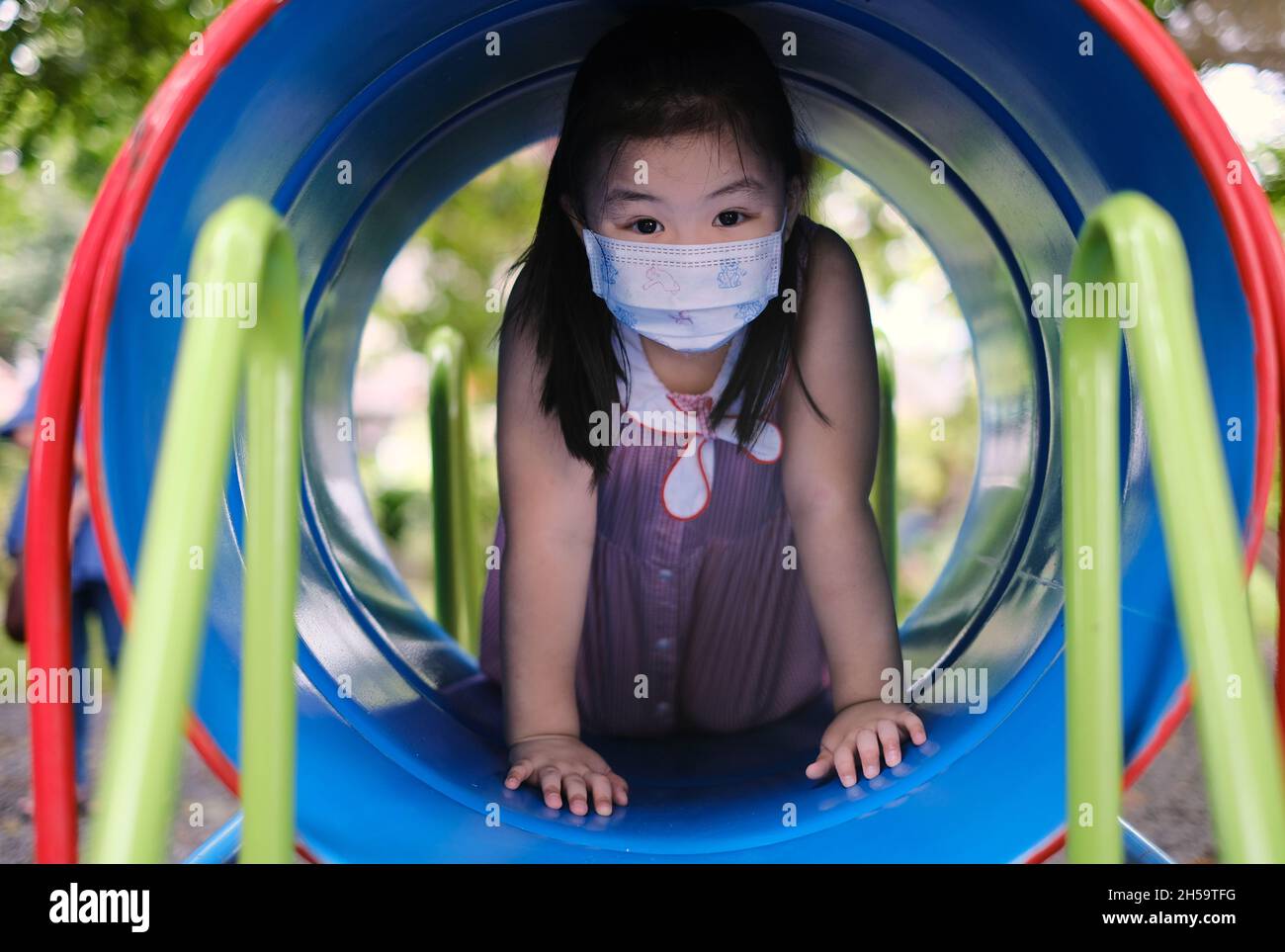 A cute young Asian girl, wearing a white mask, is playing outdoor alone, crawling through a colorful tunnel in a jungle gym of a playground in a park Stock Photo
