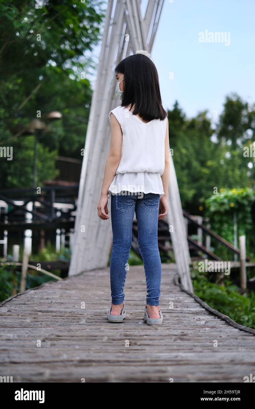 The back view of a cute young Asian girl in white shirt and blue jeans walking along a long wooden path with green plantation along the side. Stock Photo