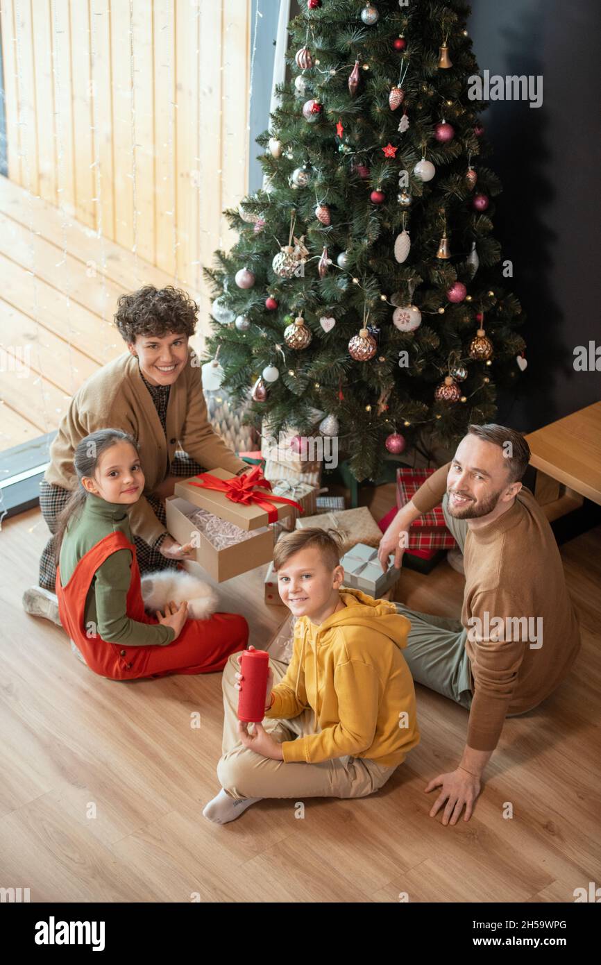 Portrait of positive beautiful Caucasian family sitting with presents on floor against Christmas tree at home Stock Photo