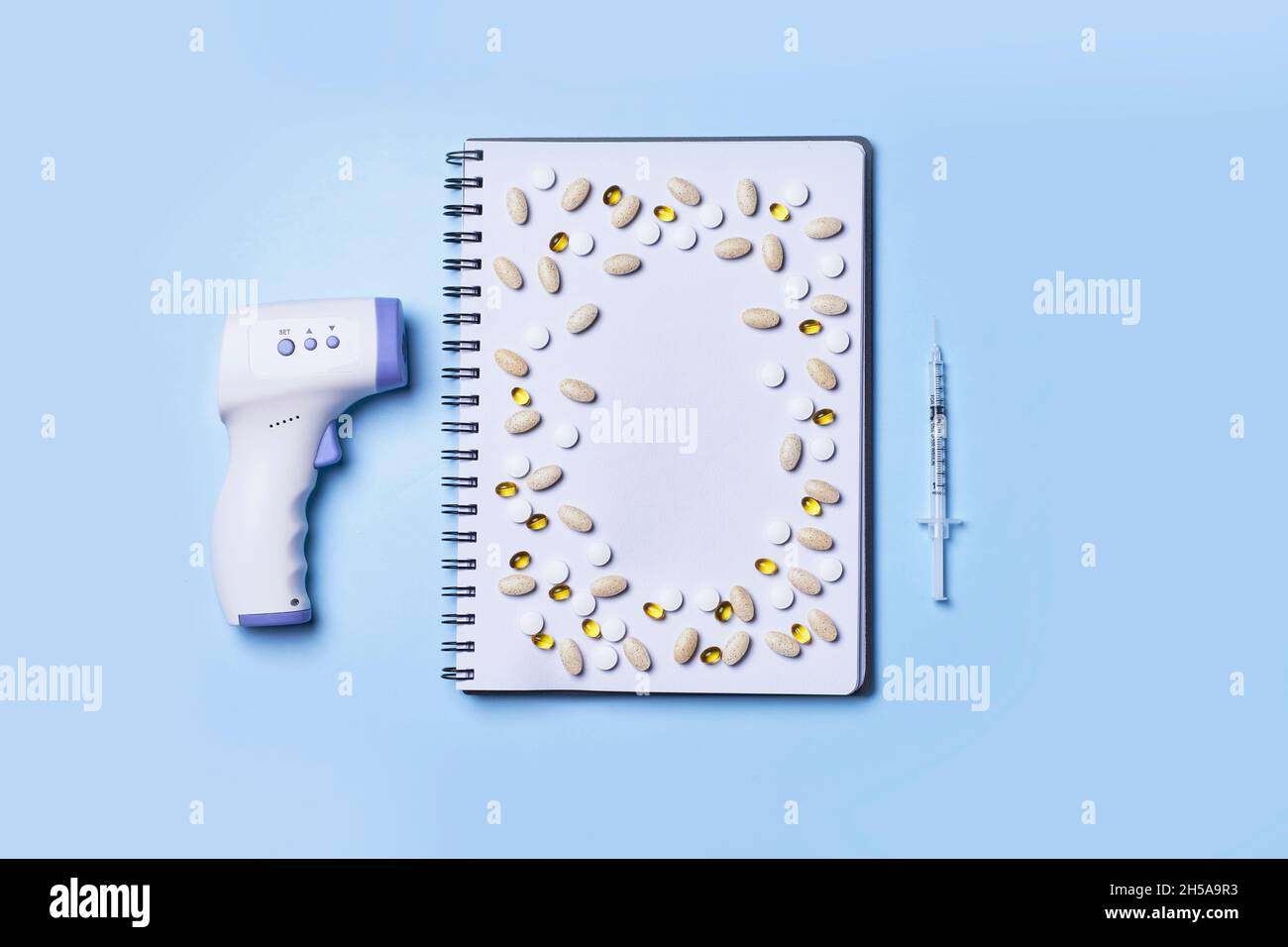 https://c8.alamy.com/comp/2H5A9R3/pile-of-medical-pills-on-notepad-with-syrine-and-infared-thermometer-on-blue-background-top-view-2H5A9R3.jpg