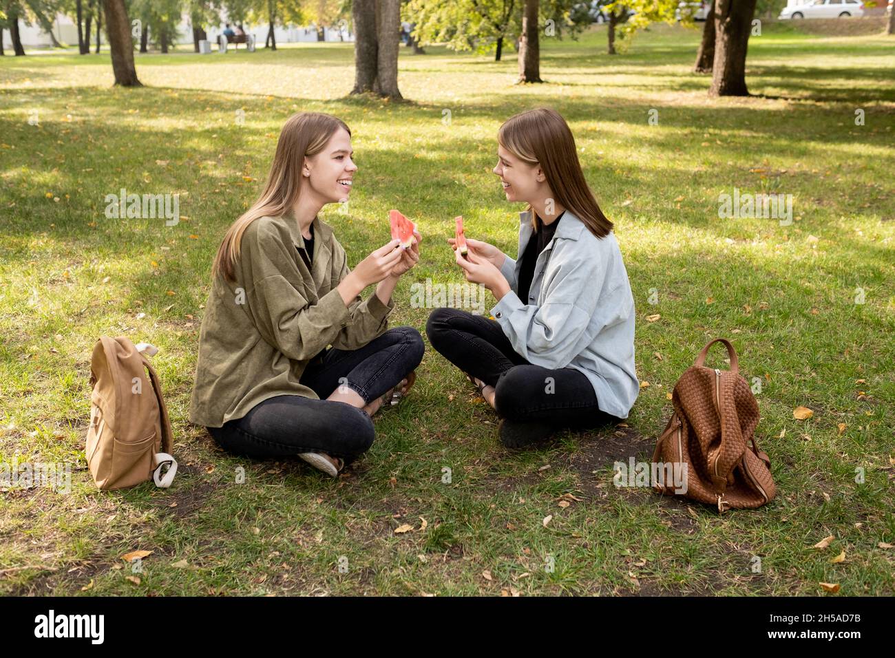 Two happy twin girls in casualwear eating sweet juicy watermelon while relaxing on green lawn in park and discussing news Stock Photo