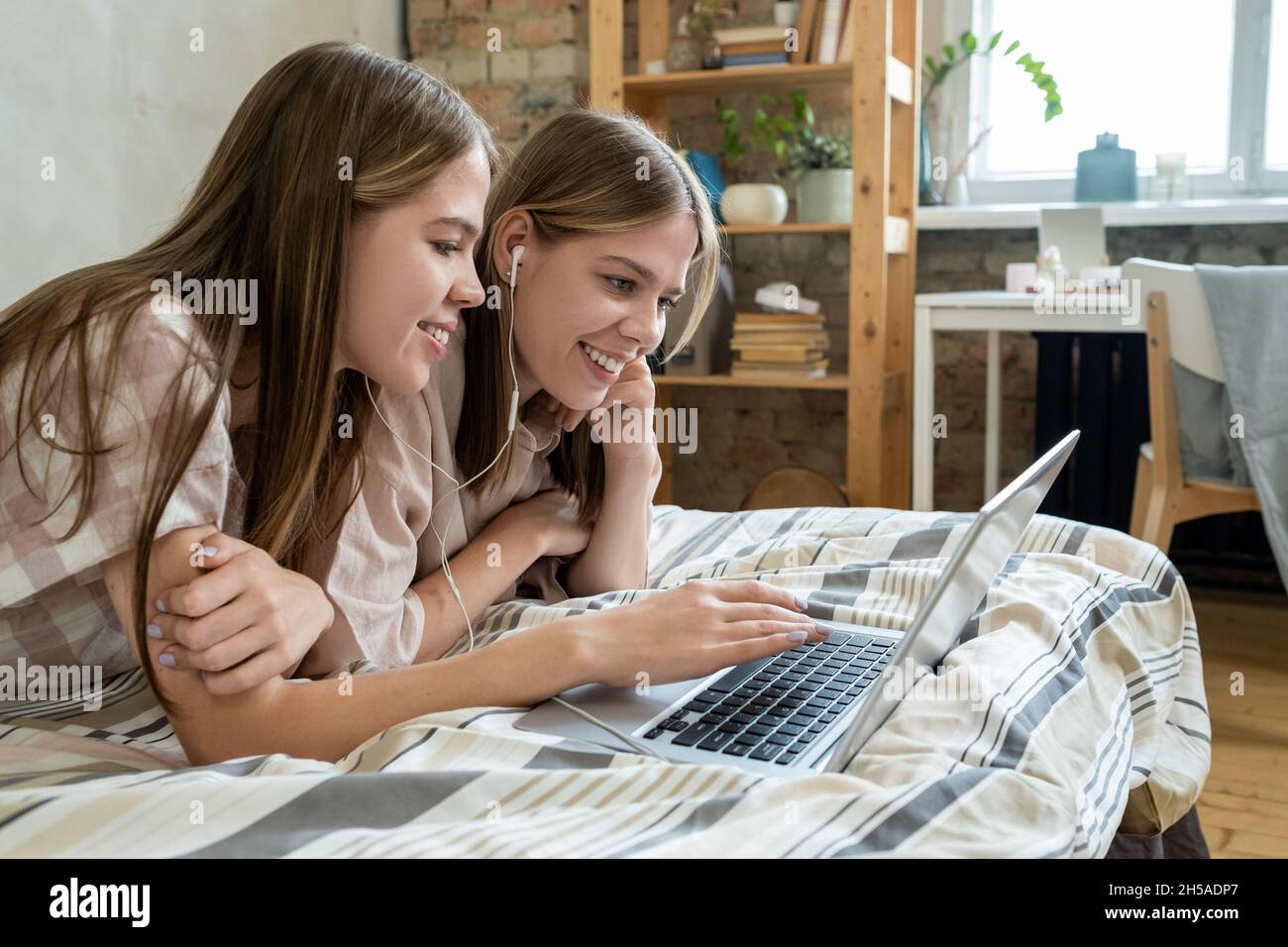Cute smiling teenage twins with earphones and laptop watching online movie while resting on bed on weekend Stock Photo