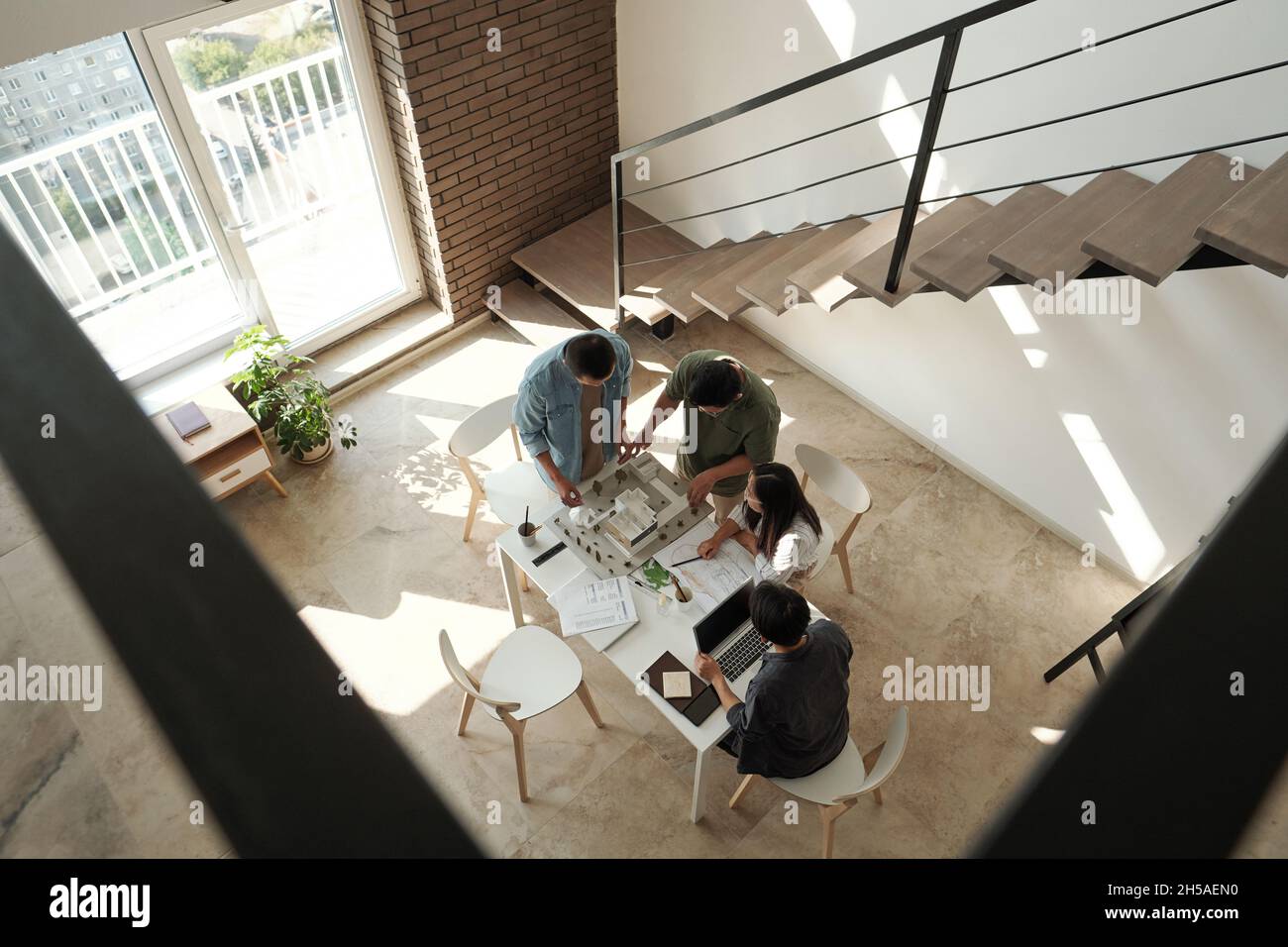 Four young Chinese architects discussing layout of new city district while standing by table in large office Stock Photo
