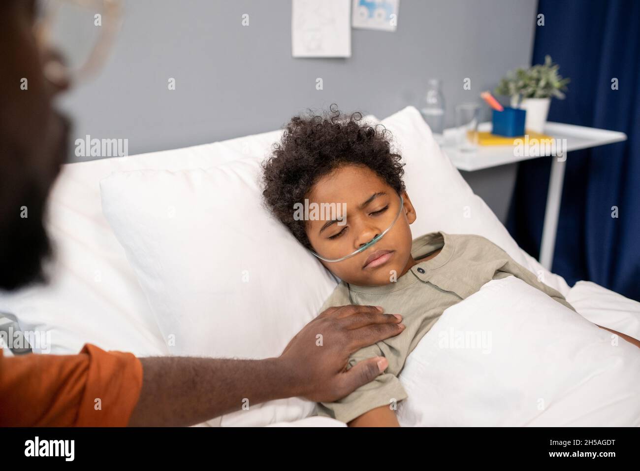 Sick little boy lying in bed with his eyes closed while father sitting near the bed and supporting him Stock Photo