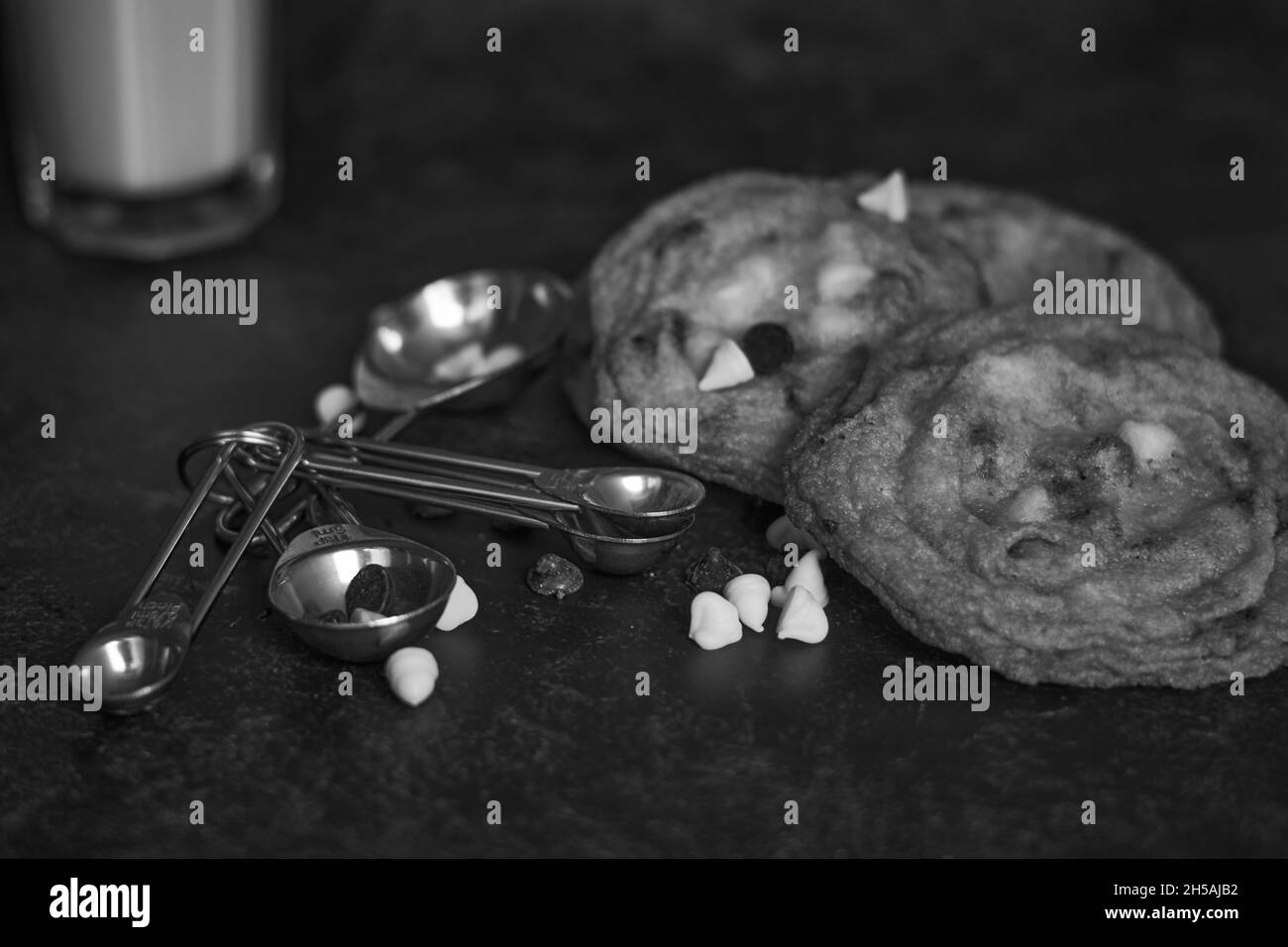 Closeup shot of chocolate chips cookies, measuring spoons, and a glass of milk in a grayscale Stock Photo