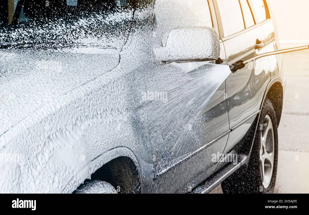 Washing car with active foam shampoo outdoors in summer in car wash station. Car exterior covered with layer of soft foam and foam spraying from hose. Stock Photo