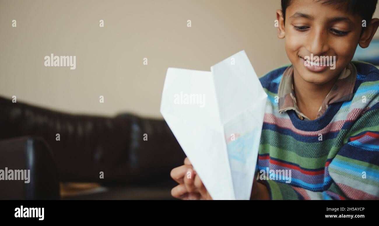 Cheerful young boy from India playing with a paper airplane at home ...