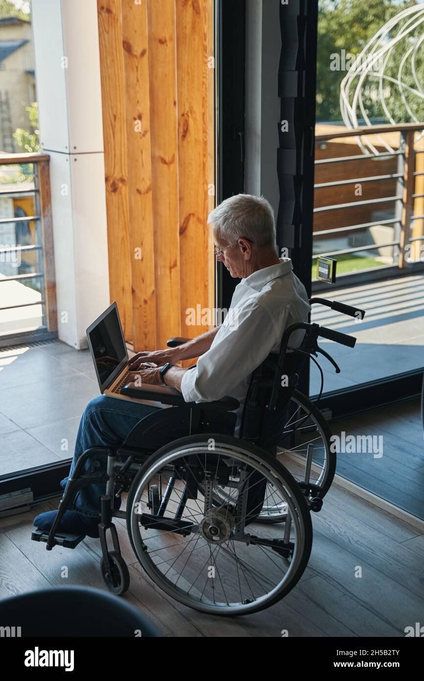 Male wheelchair user working on laptop near house window Stock Photo