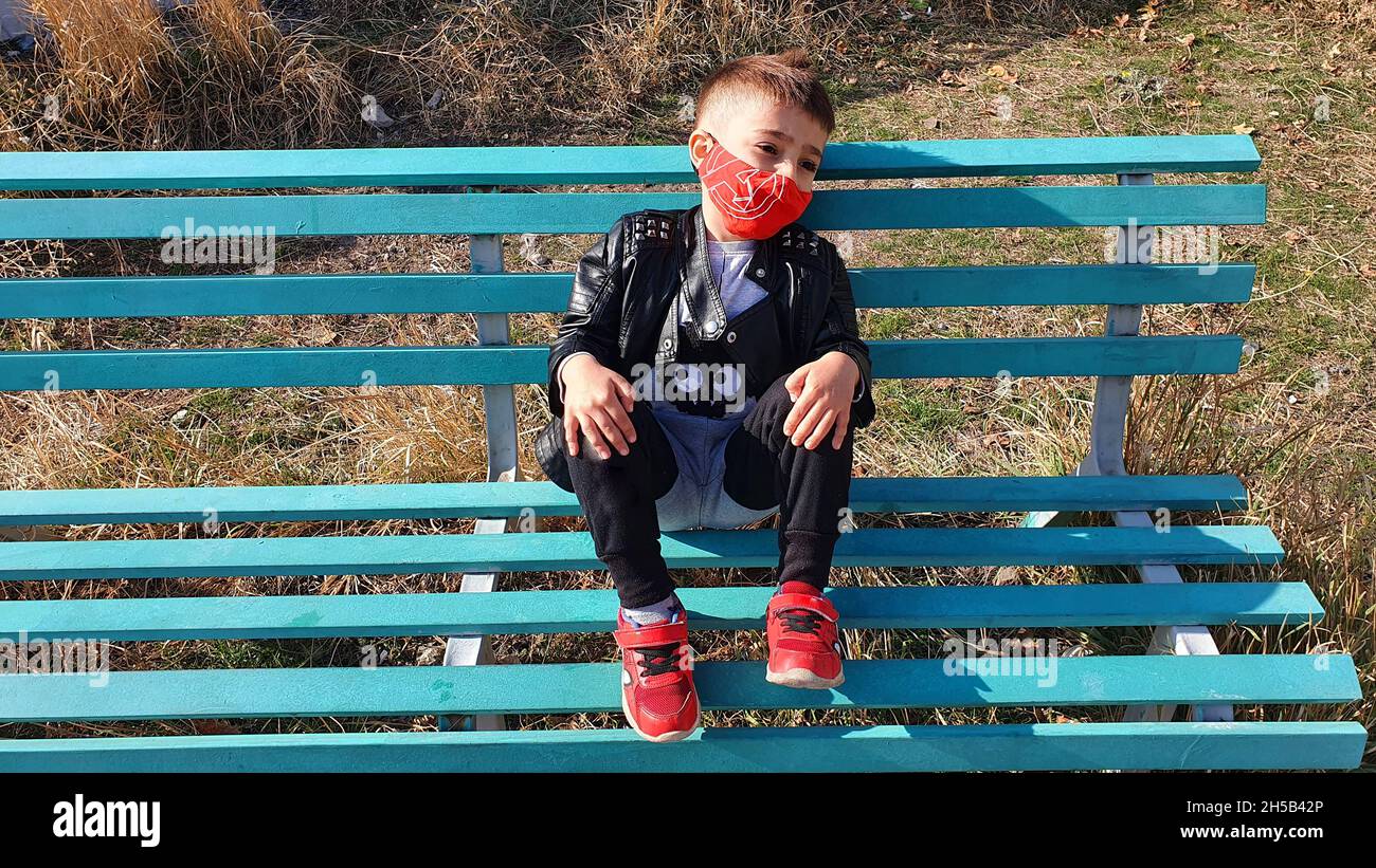 A young boy in a red medical mask sitting on a bench alone looking very ...