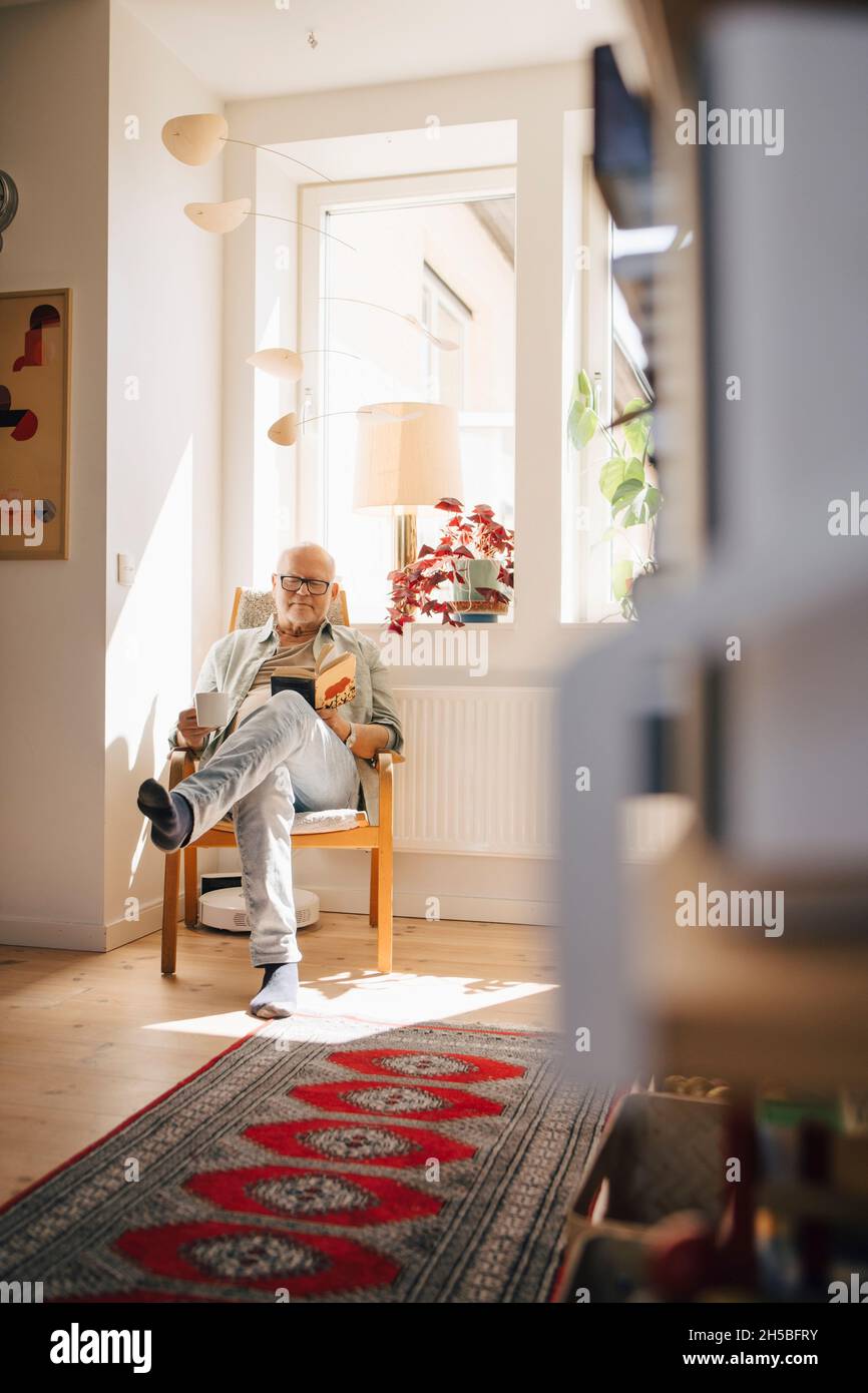 Senior man reading book while having coffee by window at home Stock Photo