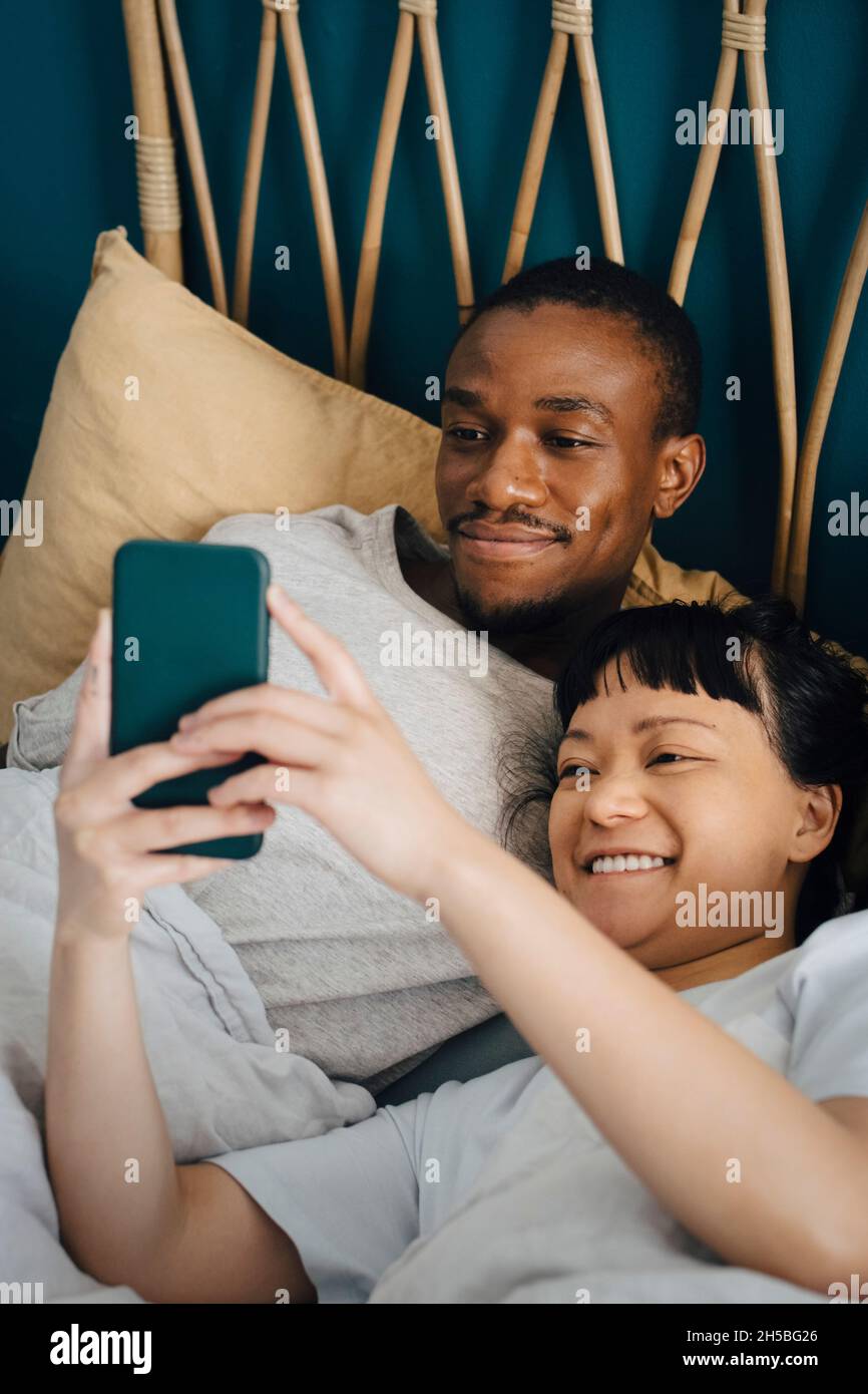 Smiling multiracial couple using mobile phone while lying on bed at home Stock Photo