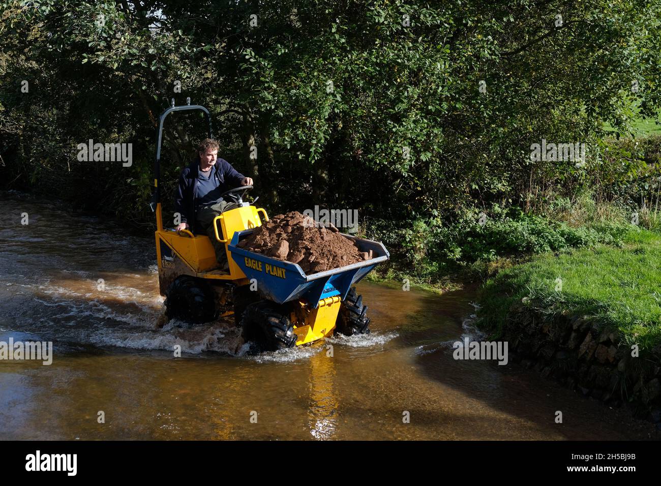 A man driving a small earth mover through water in Dunster, Somerset. Stock Photo