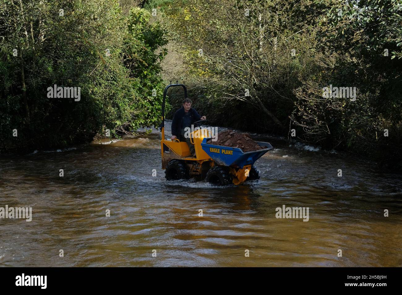 A man driving a small earth mover through water in Dunster, Somerset. Stock Photo