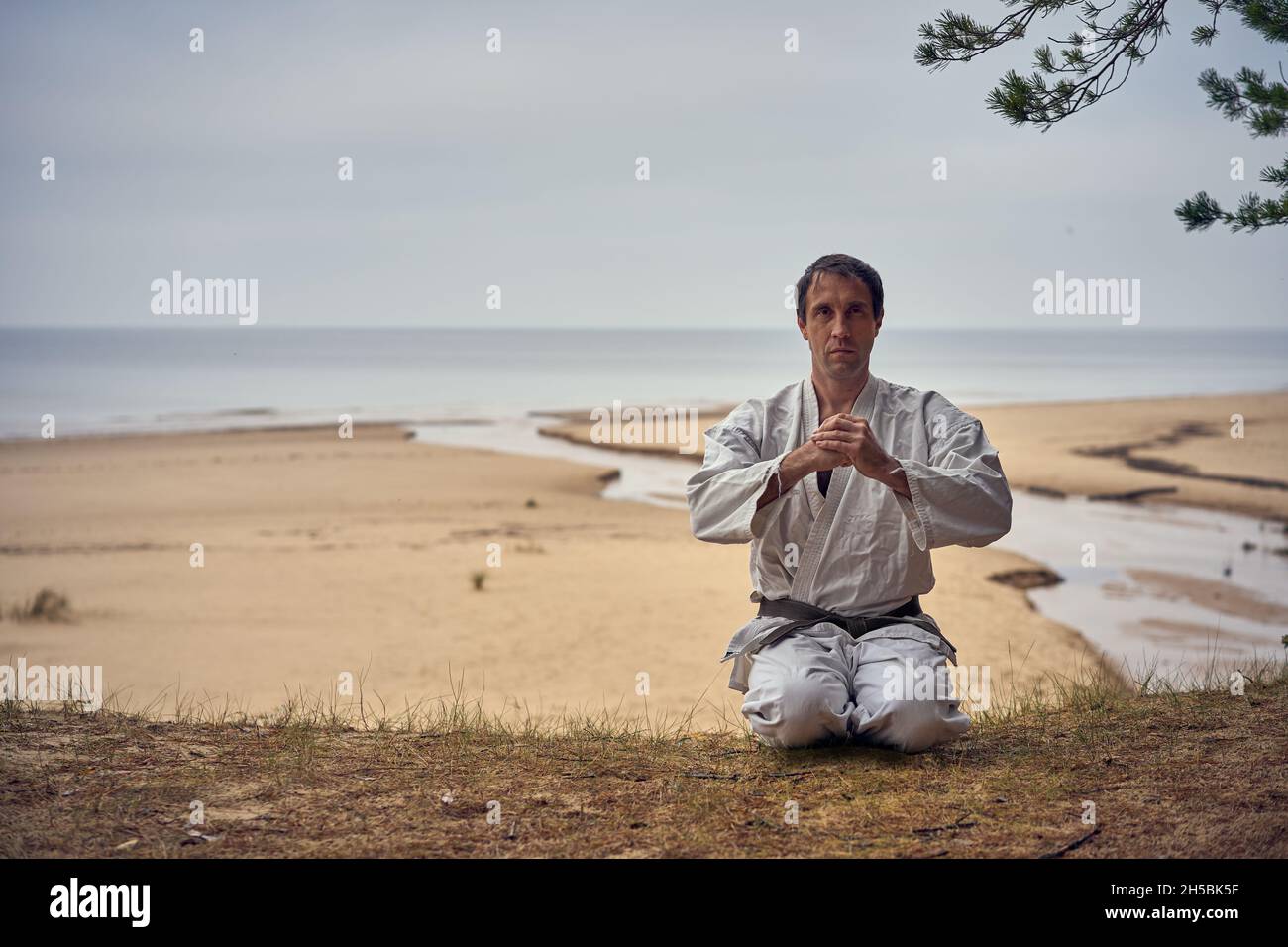 Karate man in an old kimono and black belt meditating at the sea. Martial arts concept. The river flows into the sea at the background.  Stock Photo