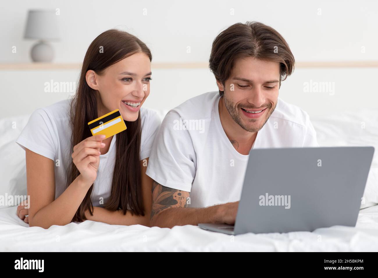 Smiling young caucasian man and lady lie on bed, paying for purchase and order with laptop and credit card Stock Photo