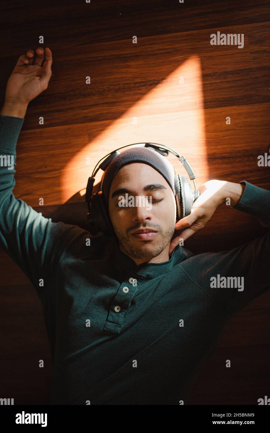 Mixed-race young millenial man laying on ground listening to music in the sunlight with headphones. Handsome hipster lying on back relaxing to music Stock Photo
