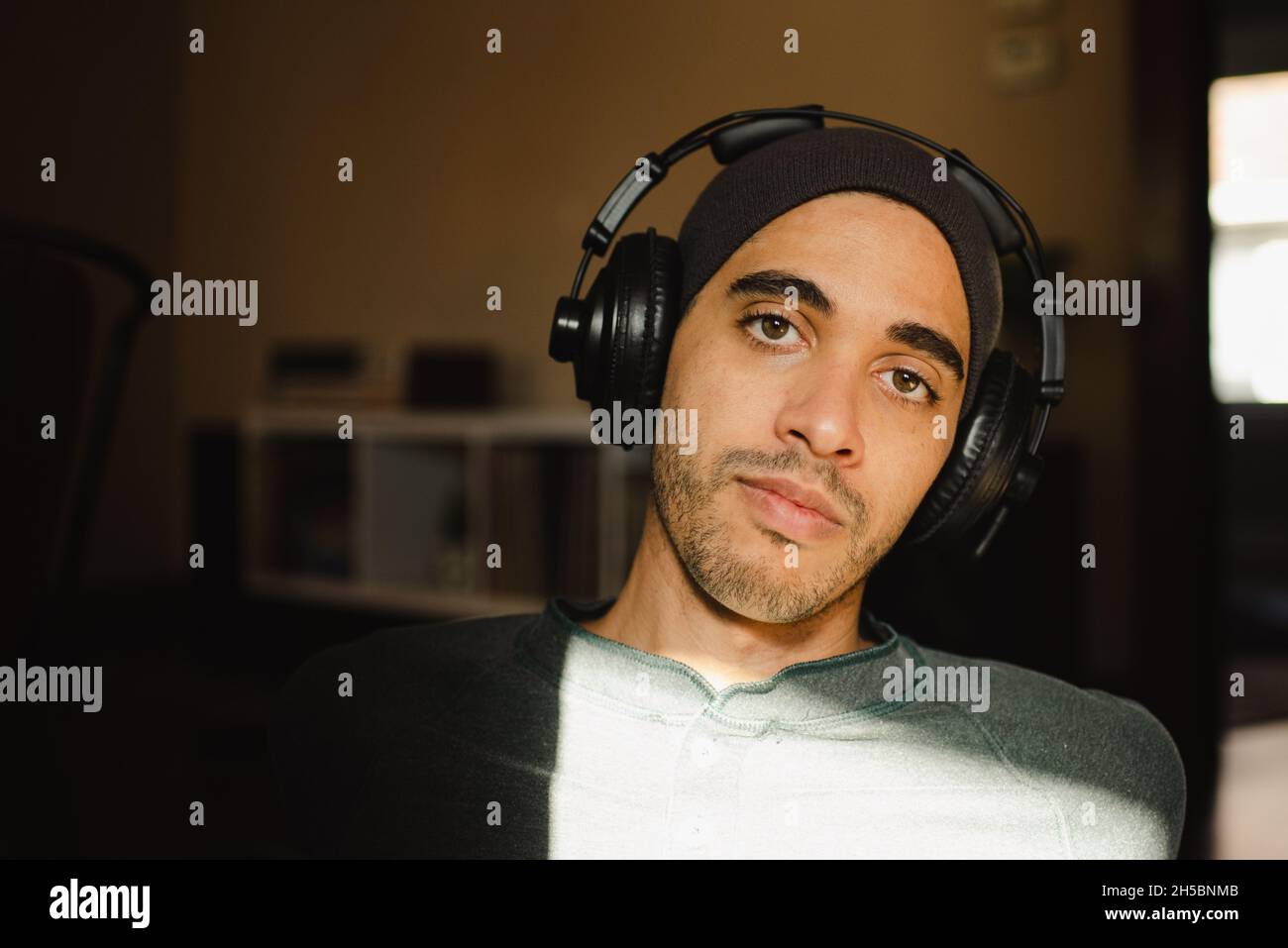 Young mixed-race hazel-eyed millennial man listening to music on over-ear headphones and looking at camera. Hipster with headphones and beanie in sun Stock Photo