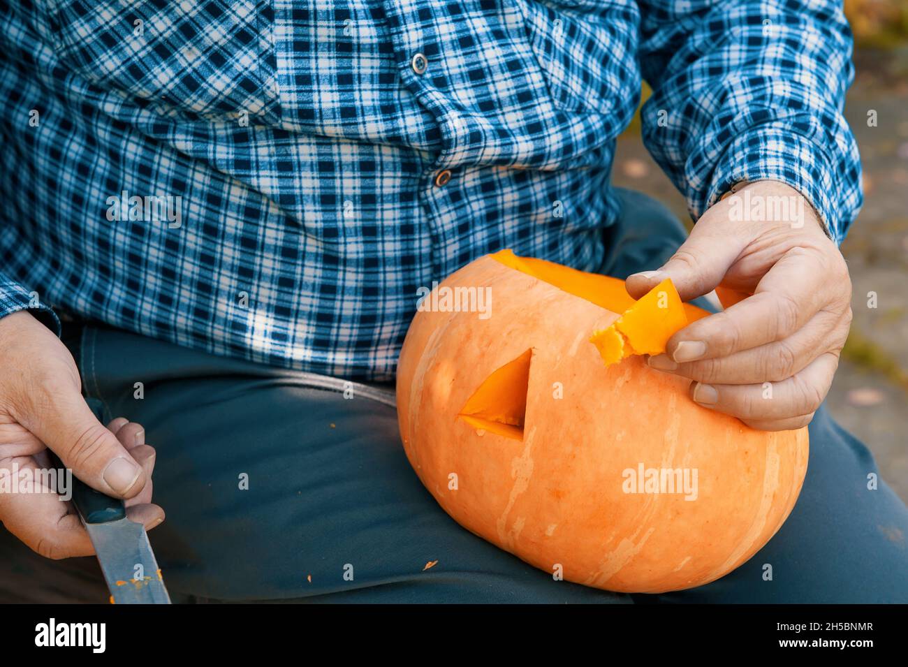 old man cuts holes in a pumpkin . a man is sitting on a bench in the garden and cooking a pumpkin for the Halloween holiday. Stock Photo