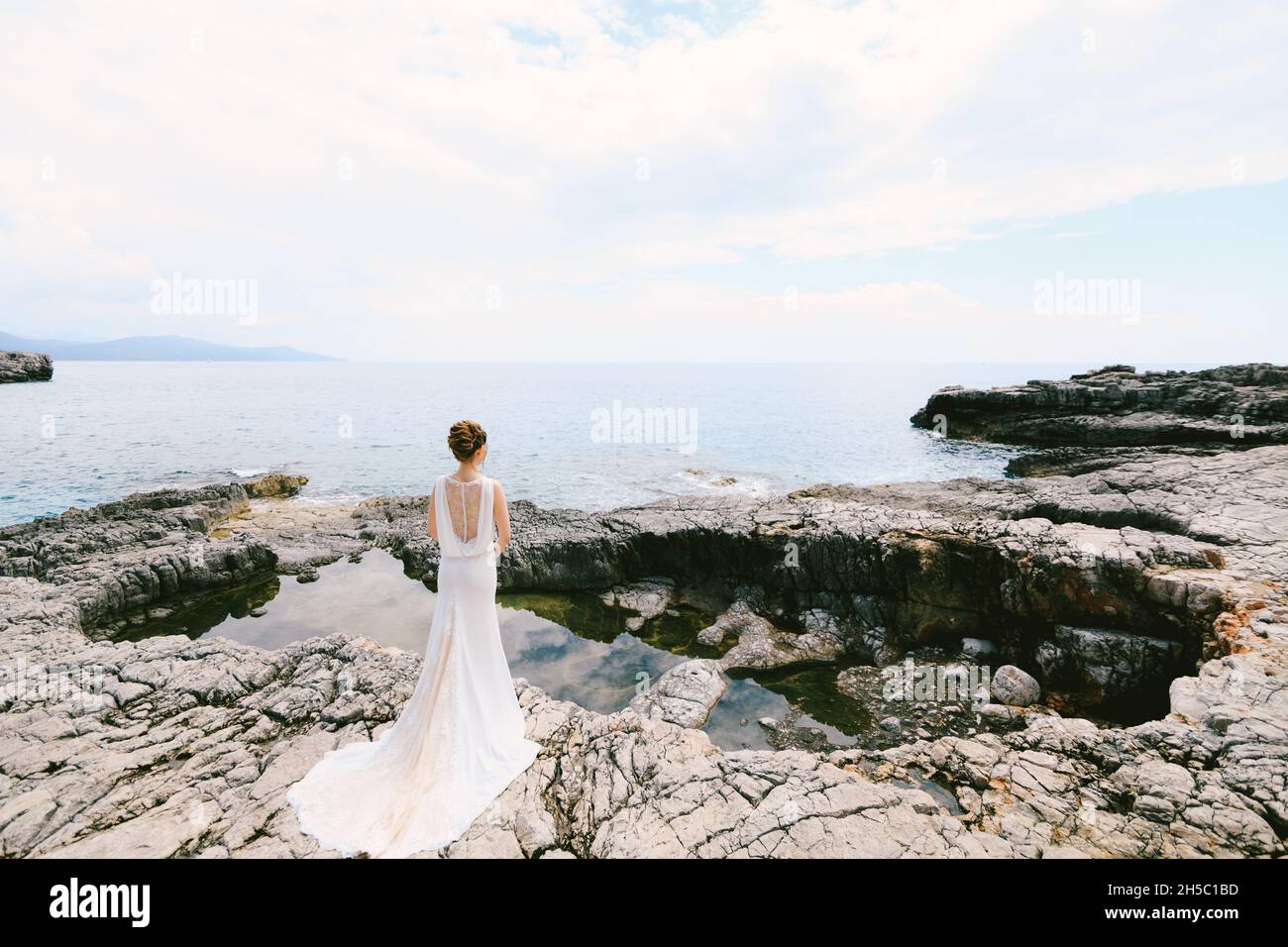 Bride in a long white dress stands on the edge of a small lake in a ...