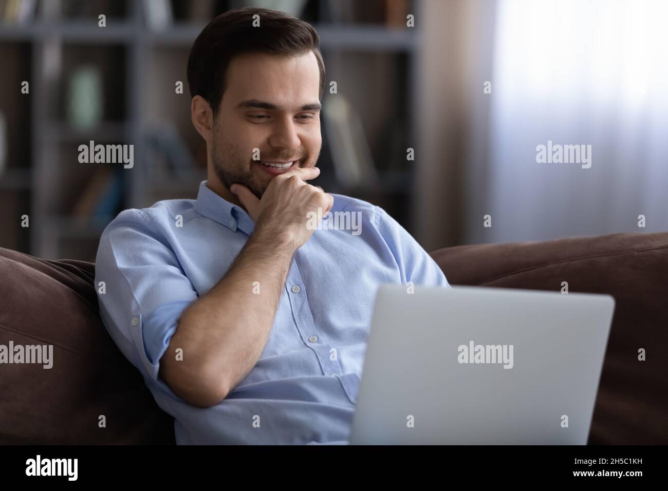 Smiling young man using computer, resting on sofa. Stock Photo
