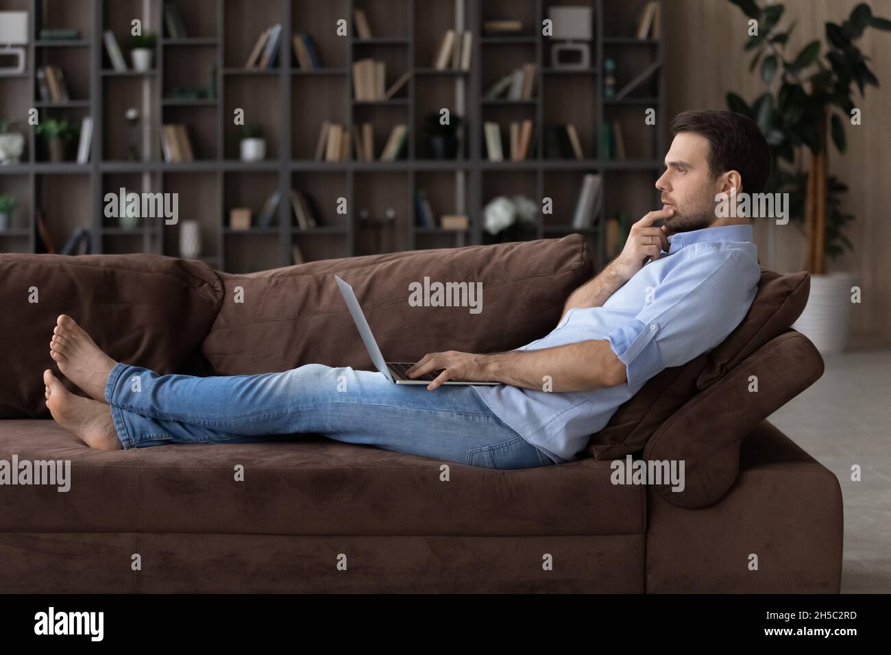 Pensive young man using computer, lying on sofa. Stock Photo