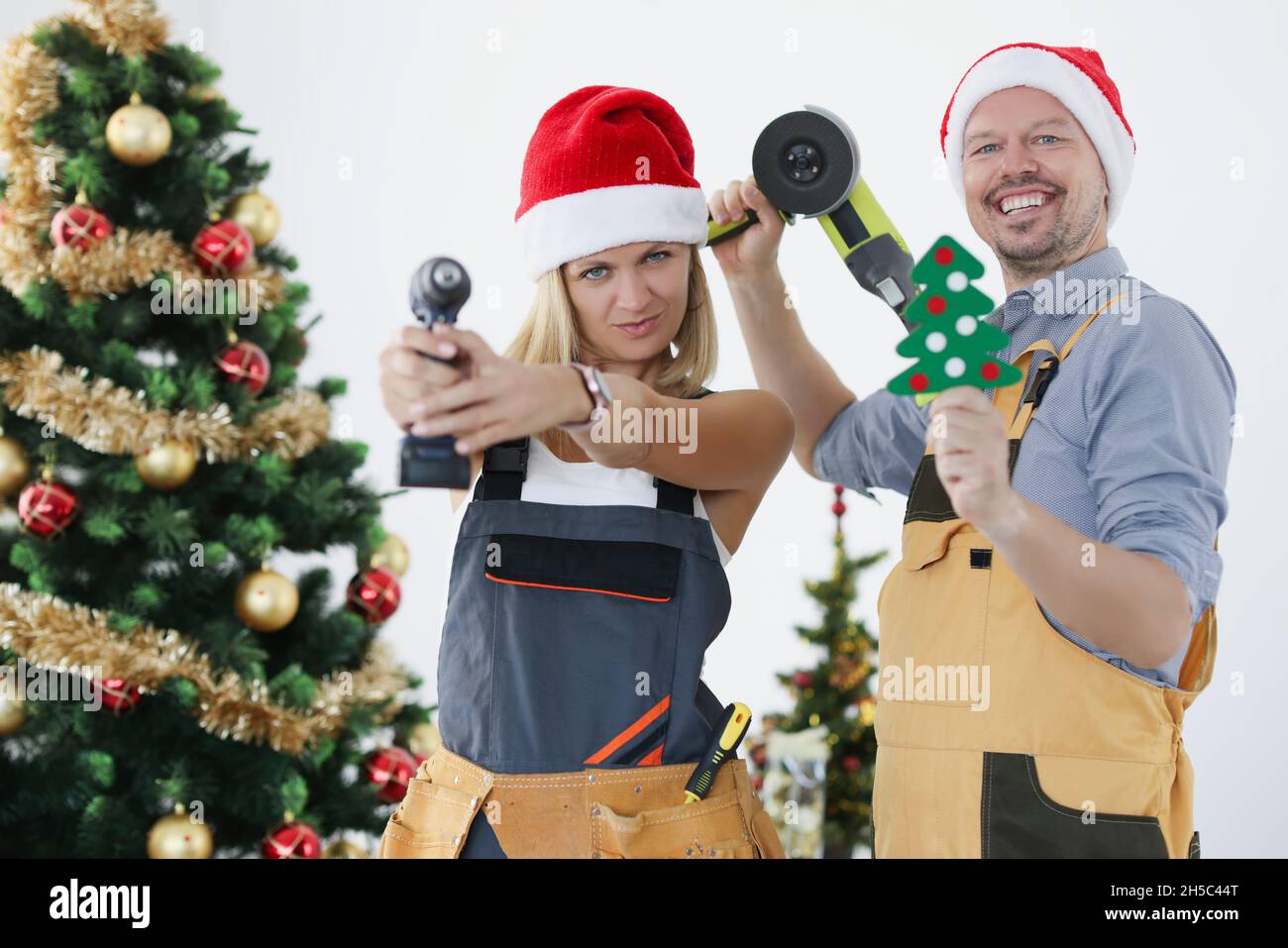 Young smiling builders woman and man in santa claus hats with tools on the background of New Year tree. Stock Photo