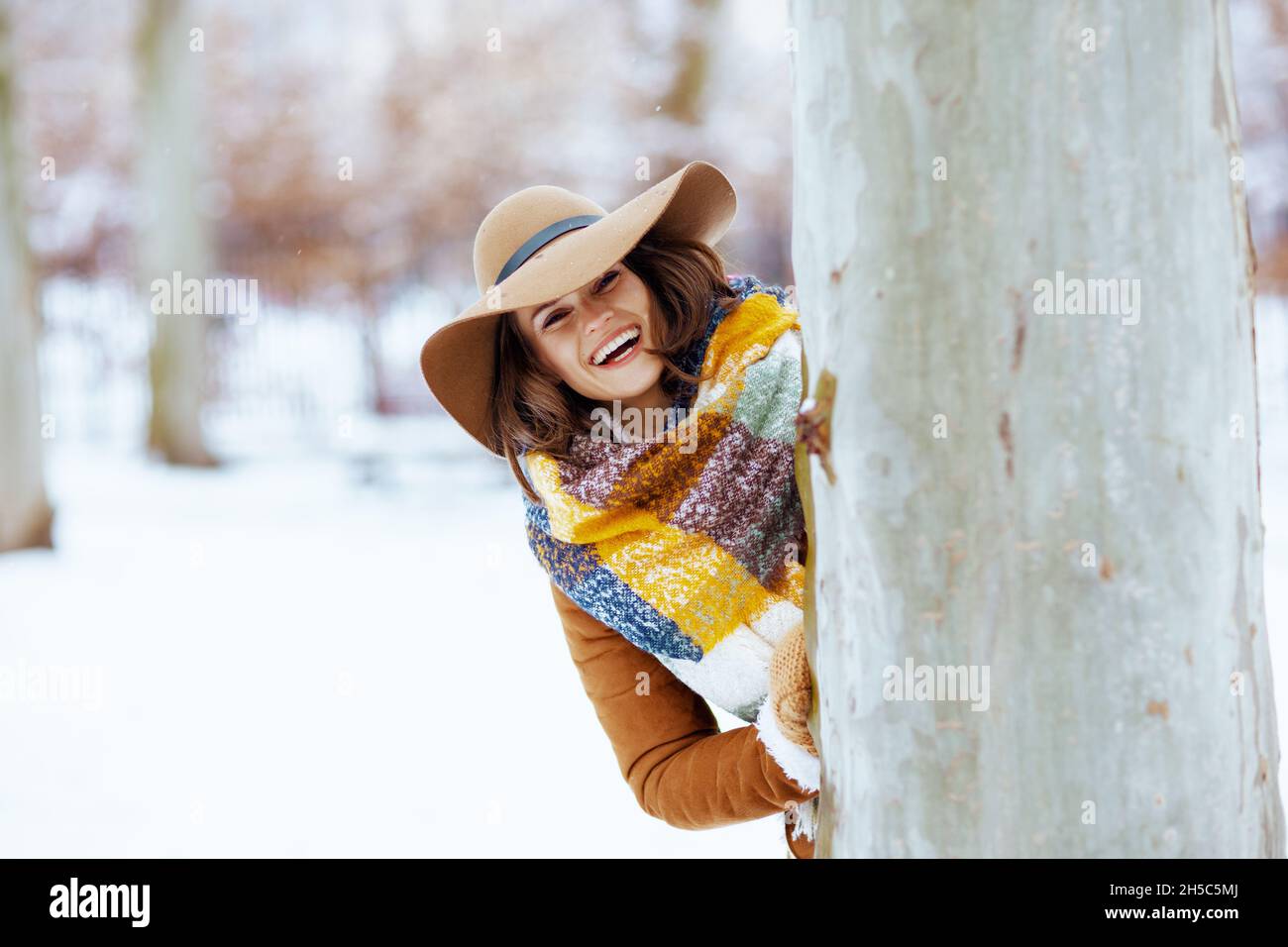 smiling stylish woman in brown hat and scarf with mittens in sheepskin coat near tree outdoors in the city park in winter. Stock Photo