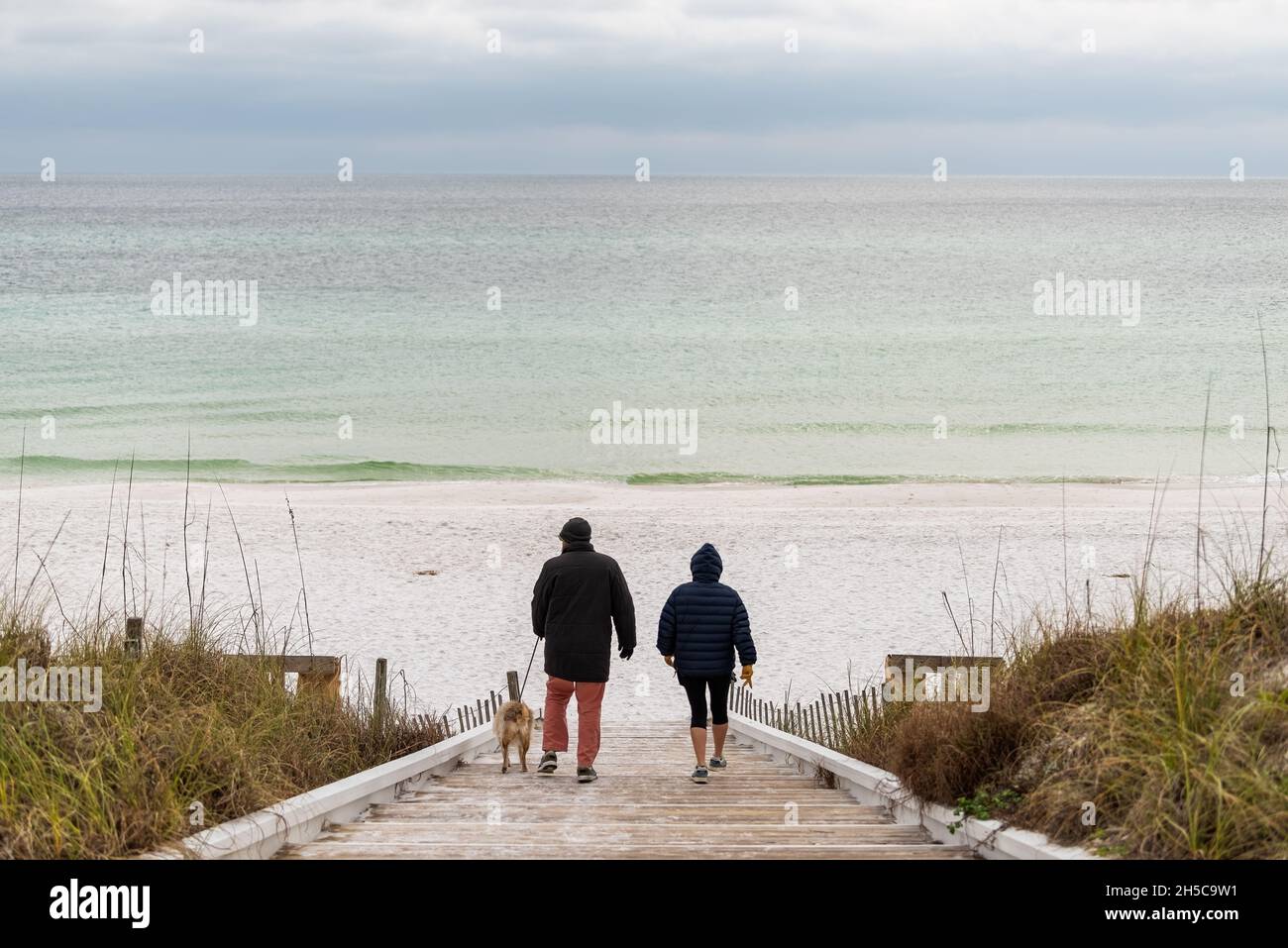 Seaside, USA - January 10, 2021: Couple people with dog walking in Seaside, Florida gulf coast beach with wooden boardwalk steps down and white sand w Stock Photo