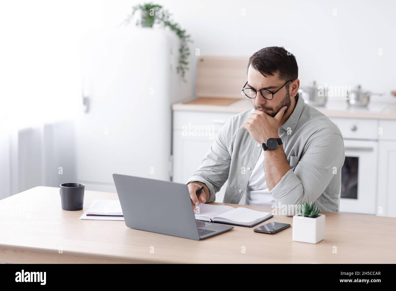Busy pensive middle aged european businessman in glasses working at computer on minimalist kitchen interior Stock Photo