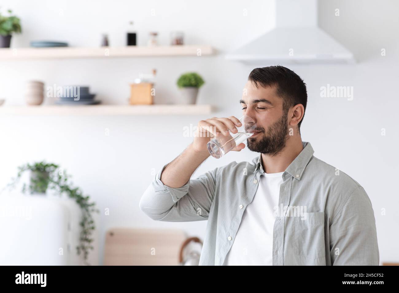 Middle aged european attractive guy with beard drinks water from glass on minimalist kitchen interior Stock Photo