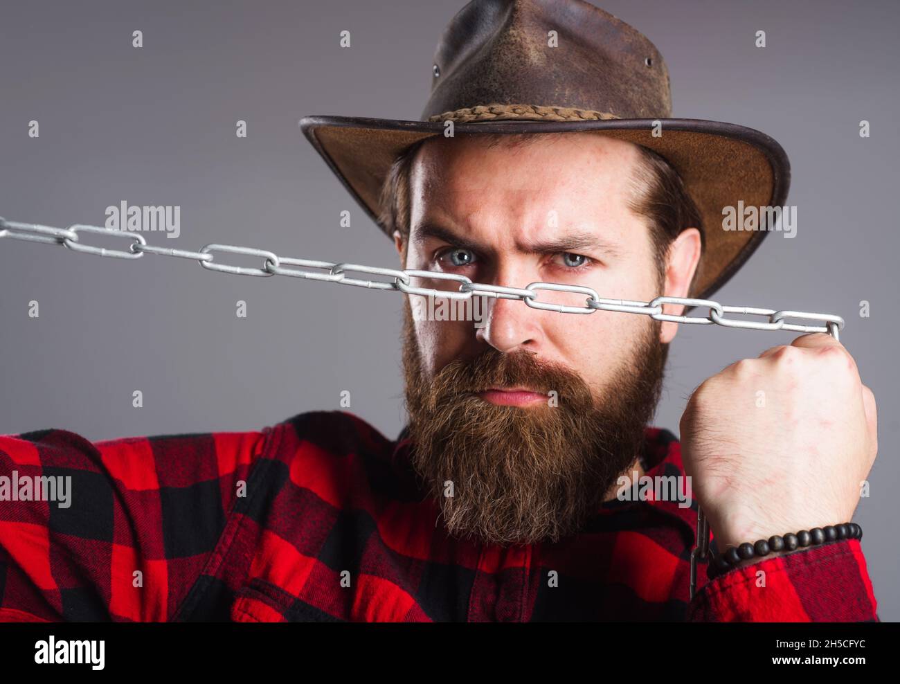 Brutal man in cowboy hat with chain. Serious bearded male in plaid shirt trying to break metal