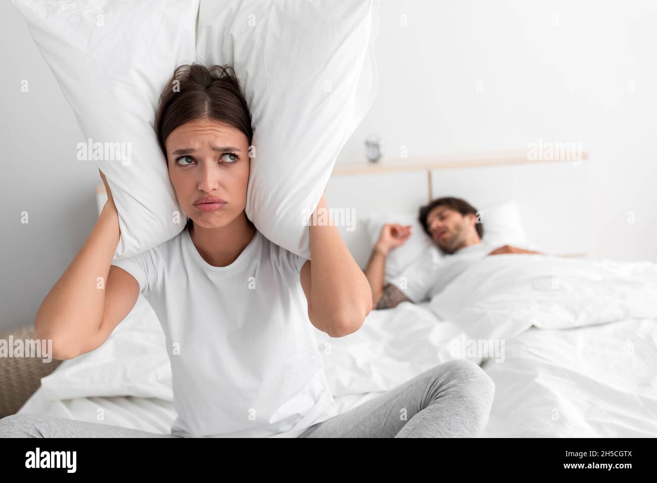 Despaired millennial european wife sits on bed covers ears with pillow and suffers from snoring