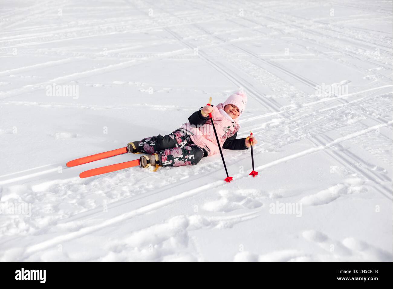 Child is lying on snow. Happy little girl in pink suit is learning to ski and fell on snowy road, snowy background Stock Photo