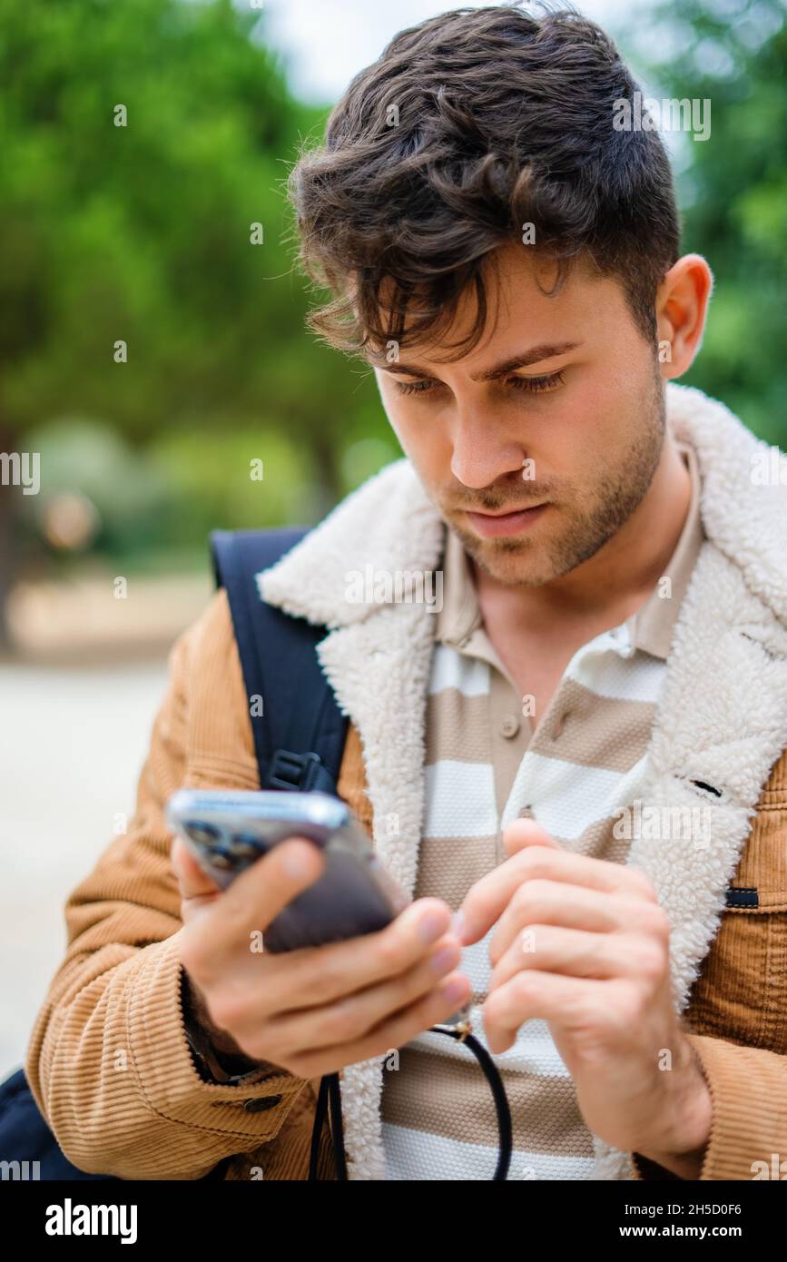 Adult male in outerwear browsing social media on cellphone while standing on blurred background of park Stock Photo