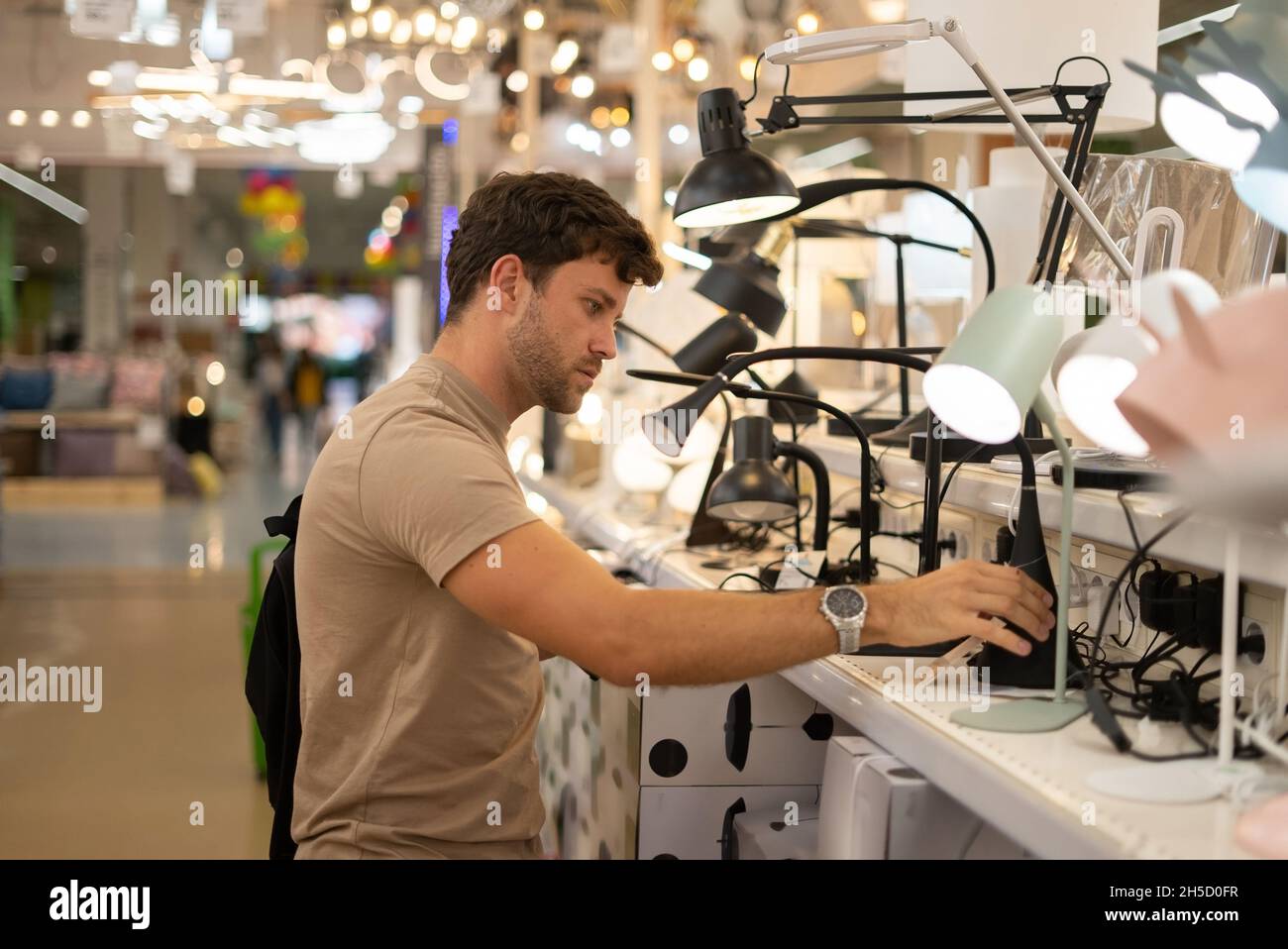 Side view of man in beige t shirt turning on and examining modern lamp while standing near shelf in shop Stock Photo