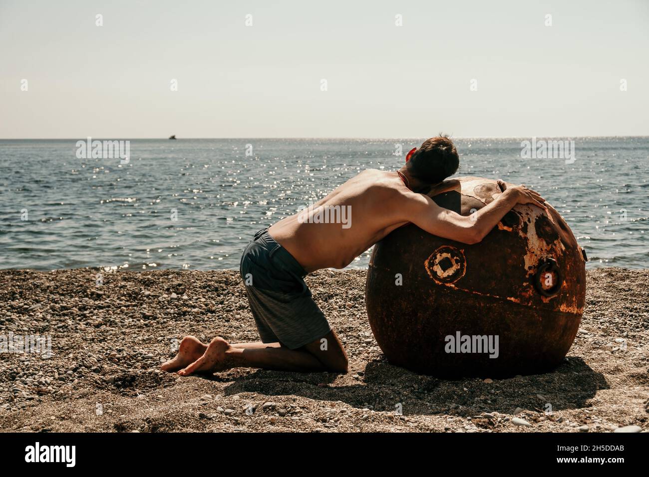 Man doing yoga and meditation outdoors near to old rusty floating marine mine on the beach with rocky shore and sea background. Healthy lifestyle Stock Photo