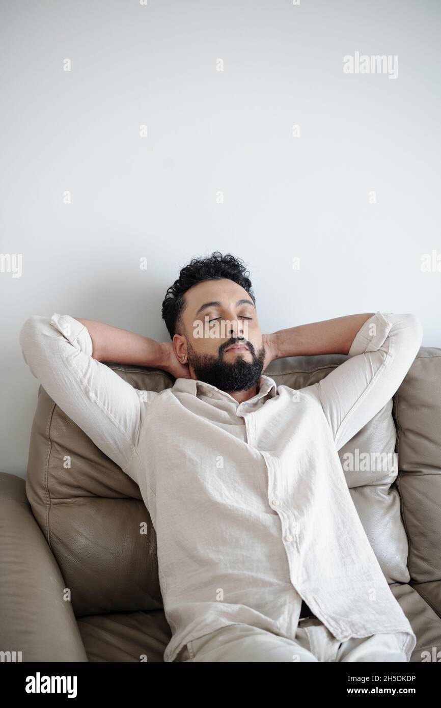 Tired man in white shirt closing eyes and leaning back on comfy sofa Stock Photo