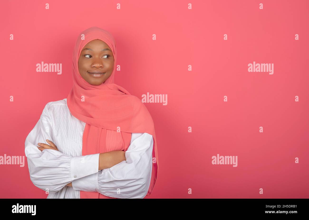 Satisfied religious female model has arms folded, looks aside, wears pink headscarf standing on pink background, copy space for text Stock Photo