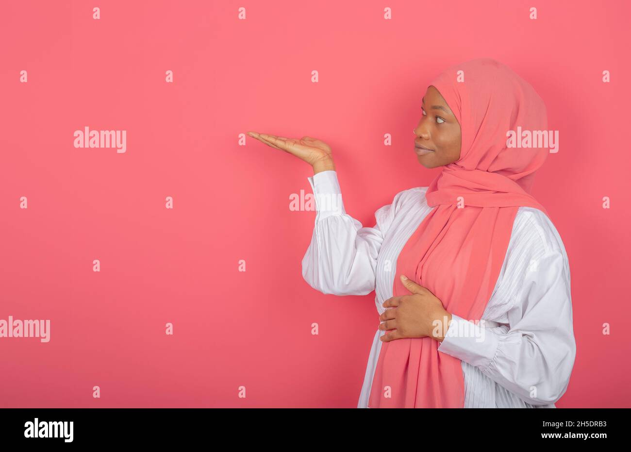 Young religious Muslim woman raises palm over blank space, holds invisible object, wears pink silk scarf on head, promotes article. Stock Photo