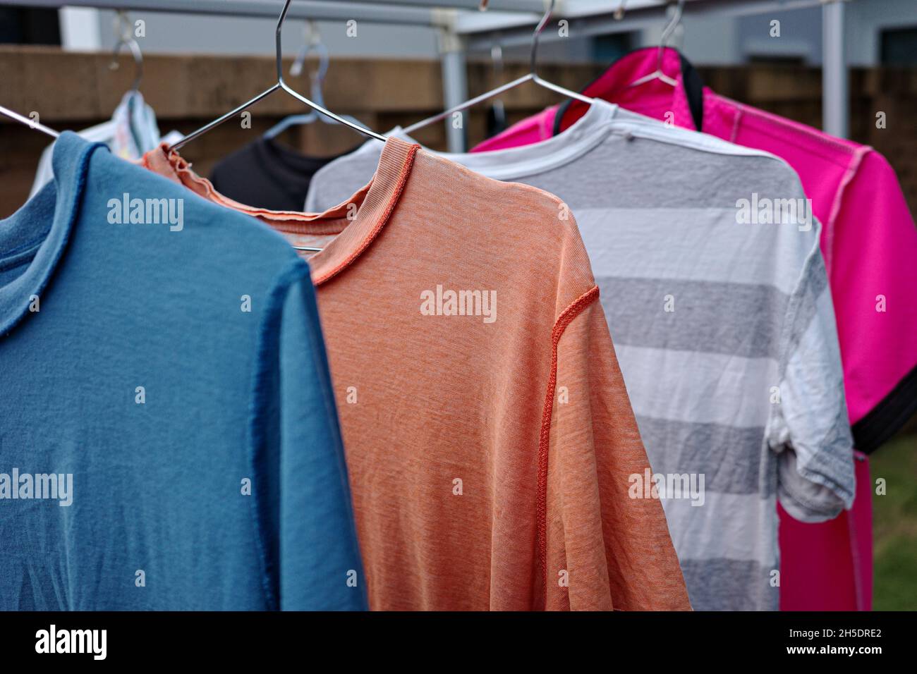 Old T-shirt in different color hanging on hanger at clothes line in outdoor with overcast sky. Stock Photo
