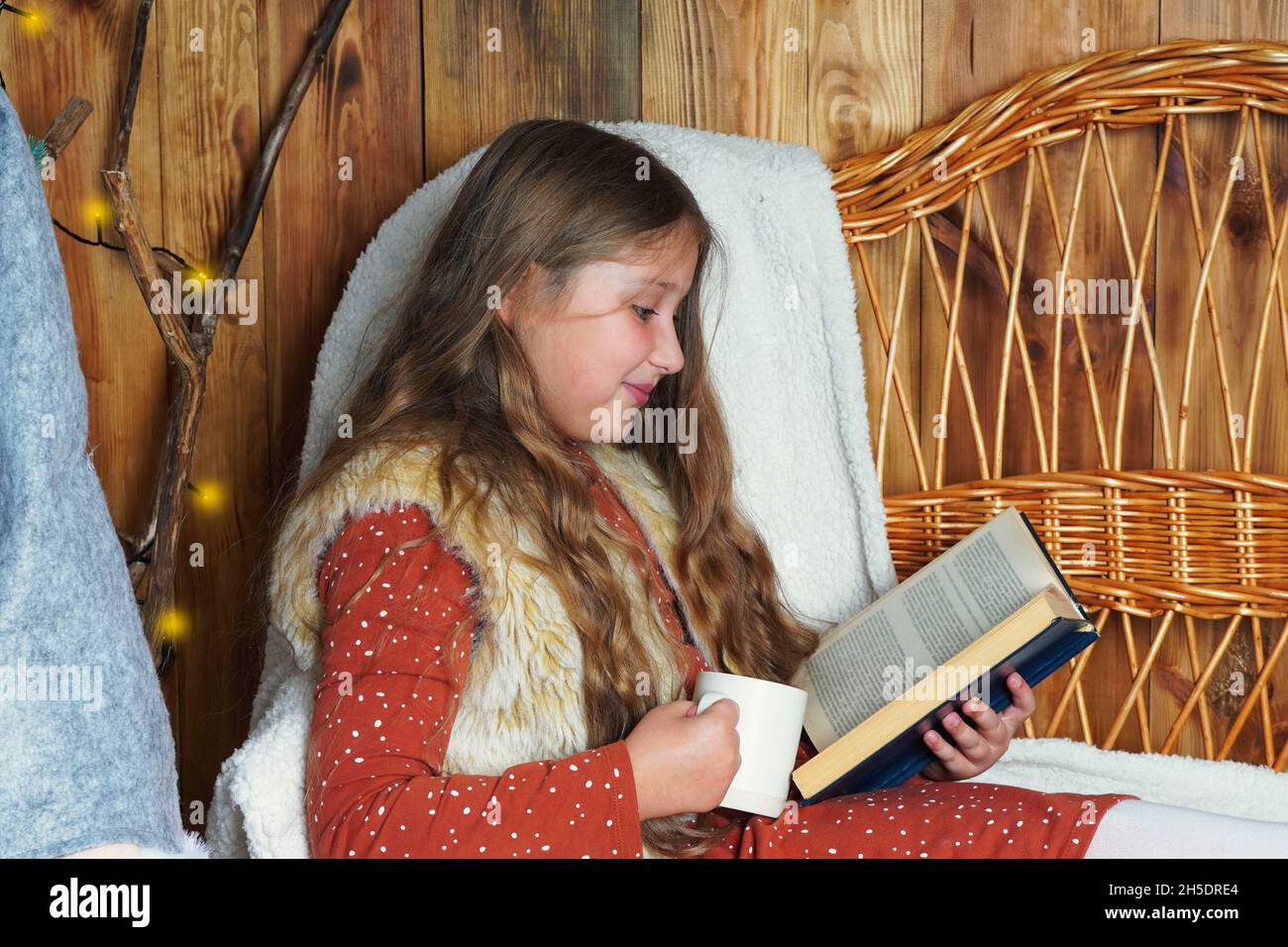 Young smiling long haired girl lying on a wicker couch, reading a book, holding a mug of hot drink near the decorated Christmas tree. Girl at Xmas. Stock Photo