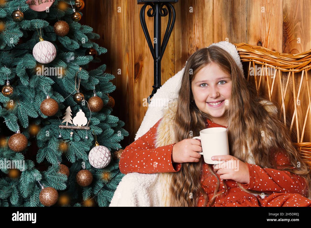 Young smiling long haired girl sitting on a wicker couch, holding a mug of hot drink near the decorated Christmas tree. Girl at Xmas. Stock Photo