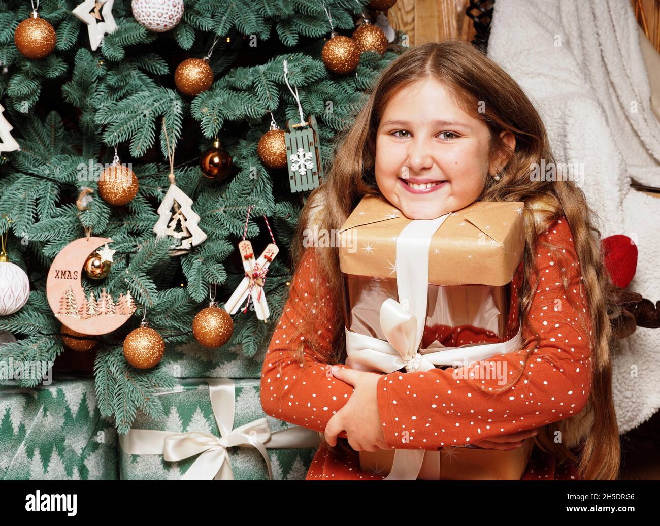 Young happy smiling long haired girl opening her Christmas gift on decorated Christmas tree background.. Girl at Xmas. Rustic concept. Stock Photo