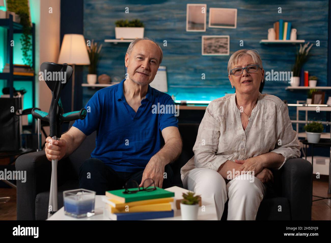 POV of married old couple sitting on living room couch together while looking at camera. Caucasian pensioners relaxing at home on sofa and enjoying retirement. Senior man holding crutches Stock Photo