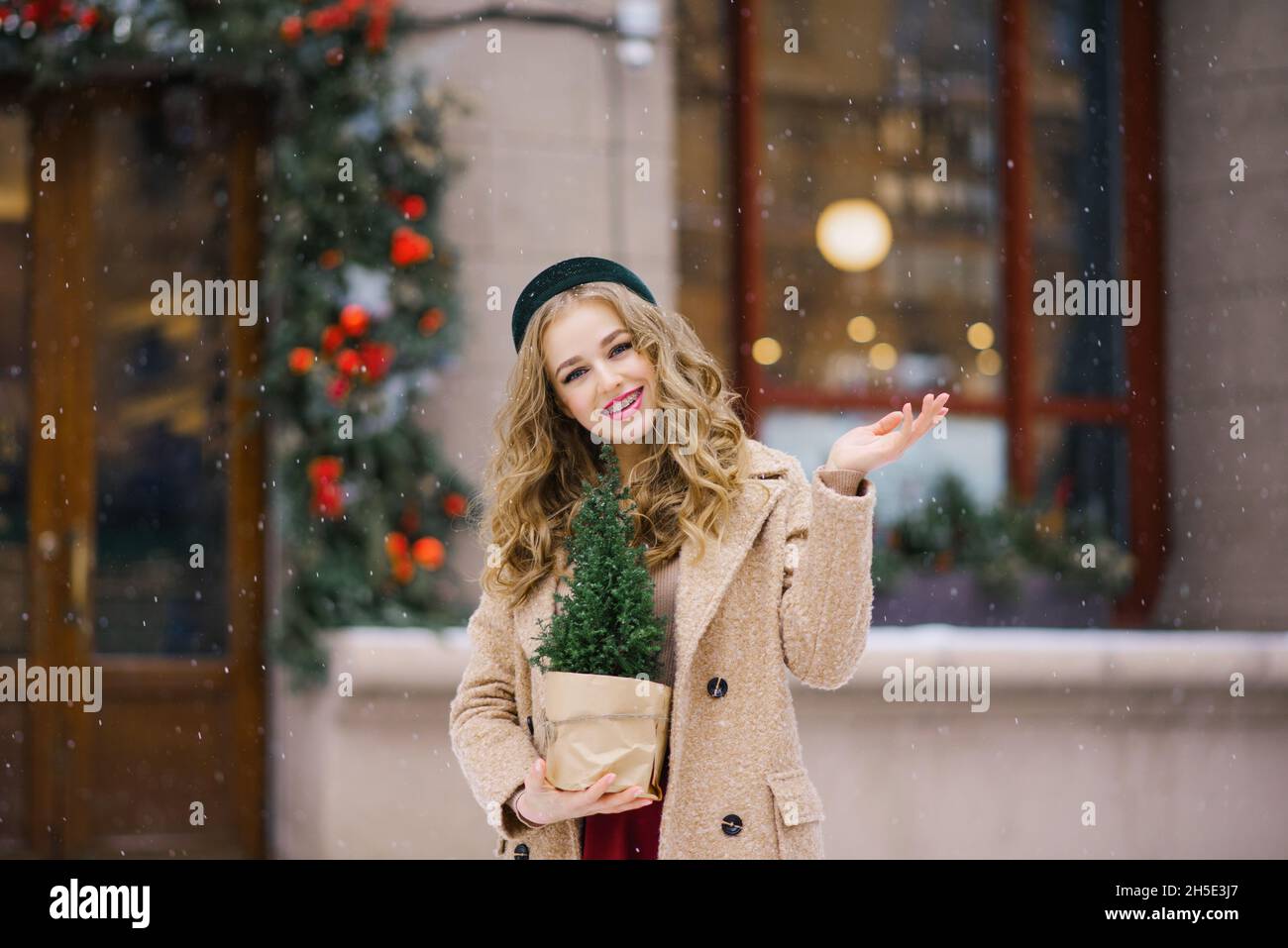 A young beautiful girl in a beige coat and a burgundy skirt is walking through a snowy city with a Christmas tree in a pot. Christmas Walk Stock Photo