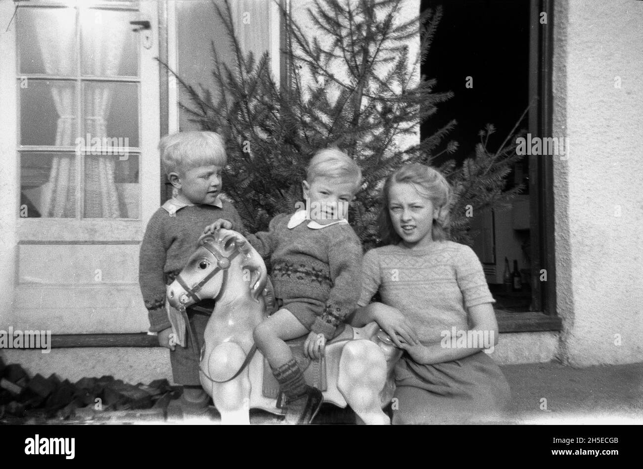 1940s, historical, outside the back of a house, a small boy sitting on a toy rocking horse, with his brother and sister beside him, England, UK, perhaps a xmas present, as there is a Christmas tree leaning against the wall by the back door. Stock Photo