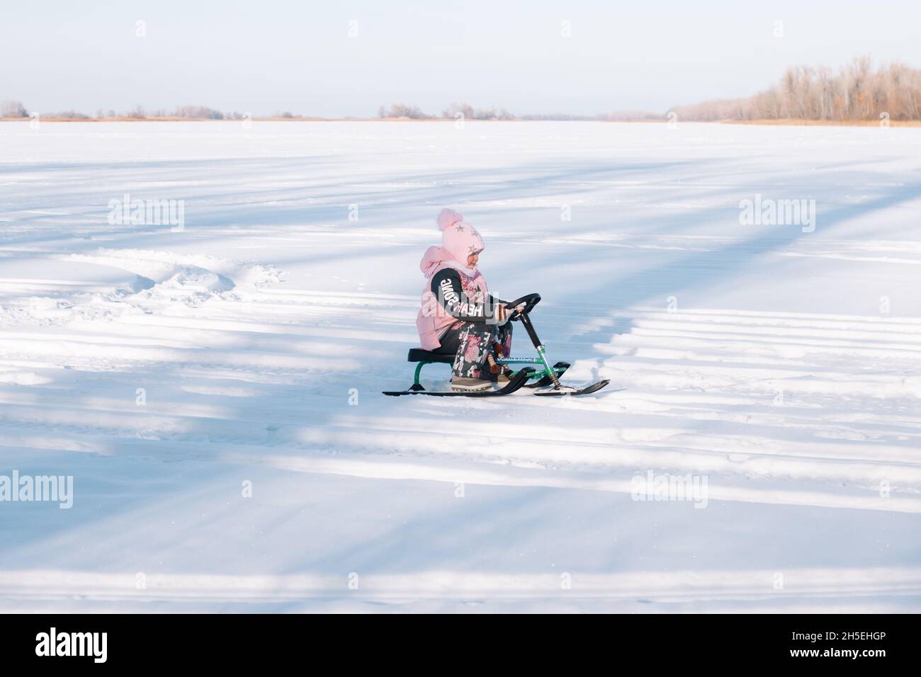 Child rides snowmobile. Little girl in pink warm jacket enjoys walk in nature and sledding on frozen river on sunny winter day Stock Photo